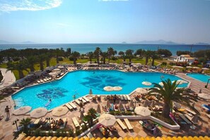 A large outdoor swimming pool with multiple sun loungers and umbrellas, surrounded by palm trees and a view of the sea. - Lagas Aegean Village (Kos Town)