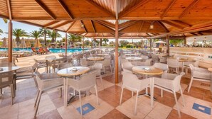 An outdoor dining area with tables and chairs, a pool, and umbrellas in the background. - Lagas Aegean Village (Kos Town)