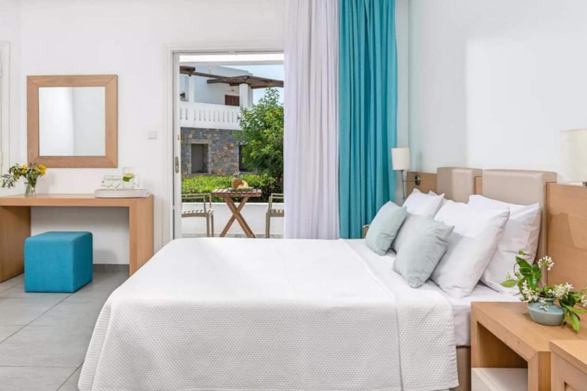 Habitación de hotel con cama, mesita de noche y escritorio con silla. Se aprecia vista de un espacio exterior con mesa y sillas.
