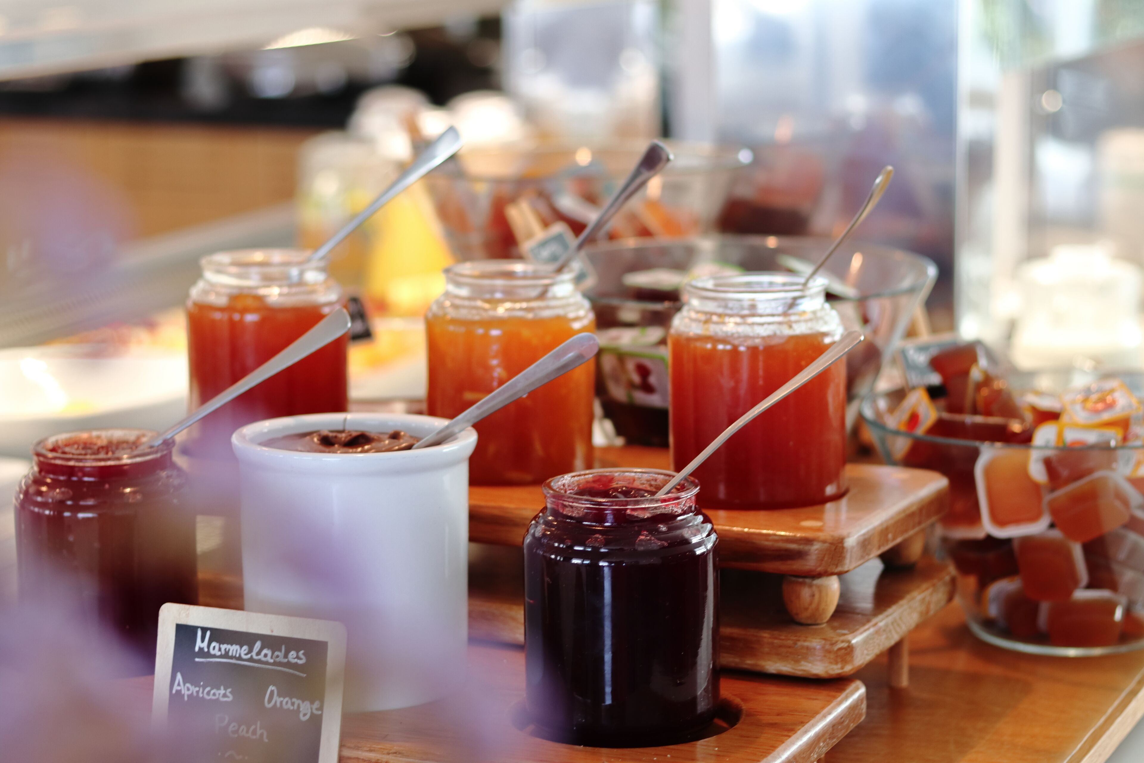 A variety of jams and jellies displayed on a wooden board with a sign indicating flavors like apricots, orange, and peach.