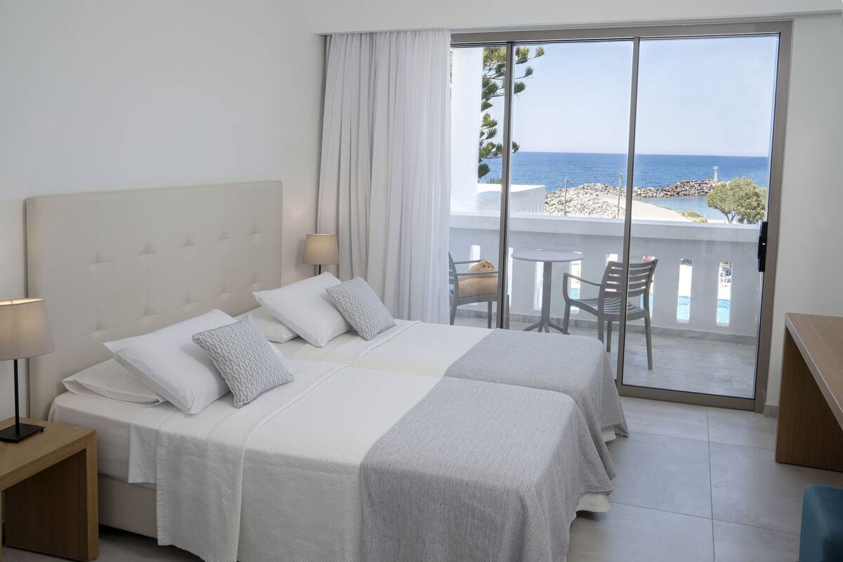Habitación de hotel con una cama grande, una mesita de noche con lámpara y vista al mar a través de una puerta corrediza de vidrio.