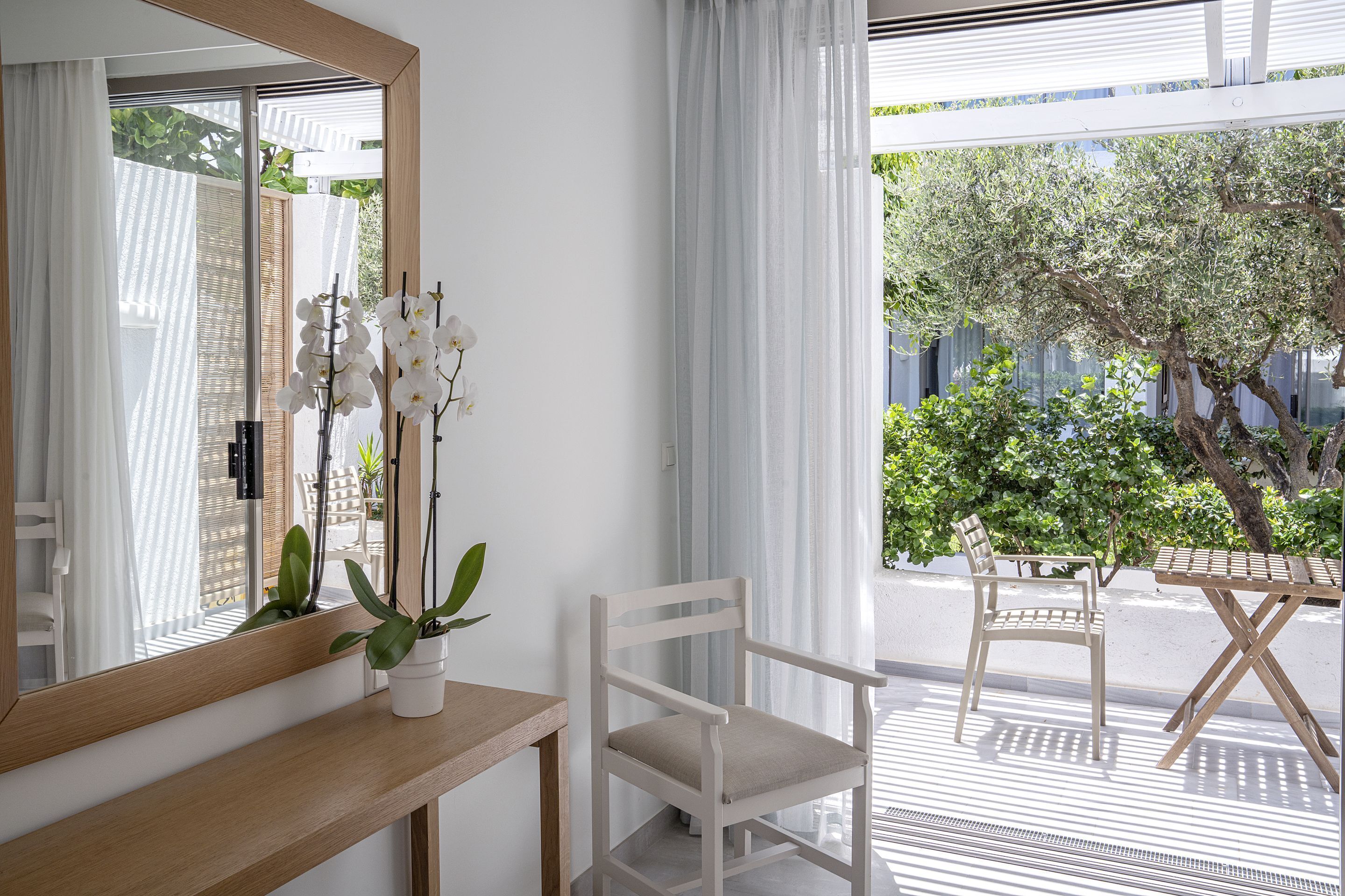 A room with a wooden desk, a white chair, a large mirror, and a view of a balcony with outdoor furniture and greenery.