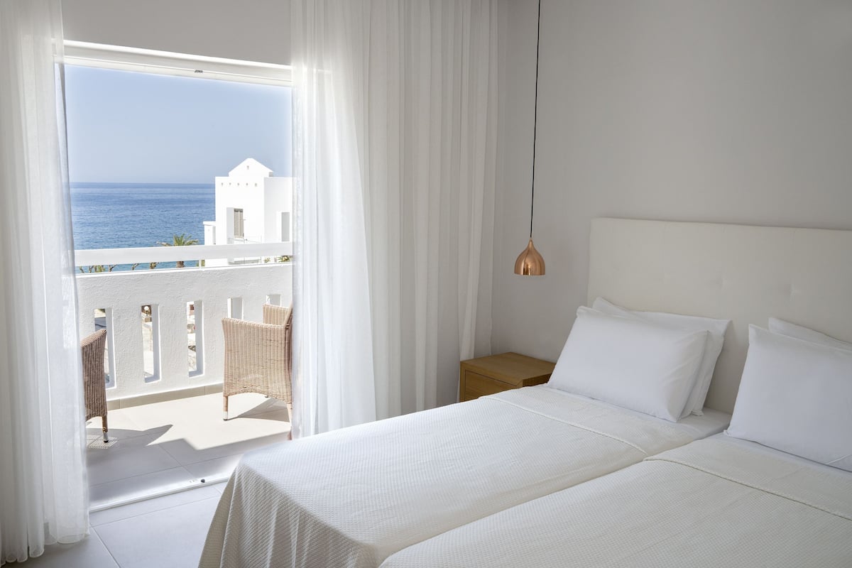 Un dormitorio con cama, cortinas blancas, vistas al mar y un balcón con sillas de mimbre.