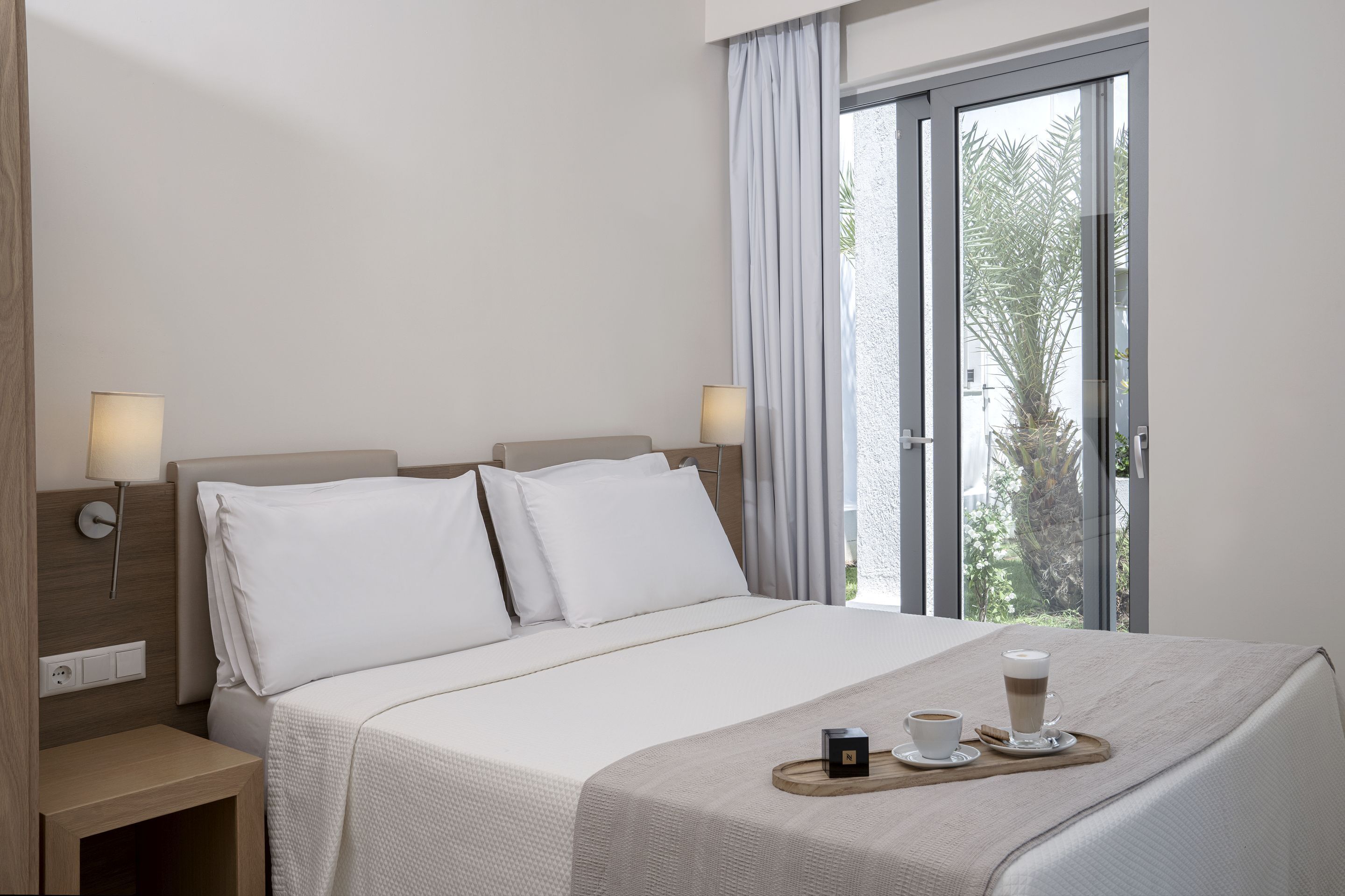 A hotel room with a bed, bedside lamps, a tray with a cup and a small box, and a window with curtains.