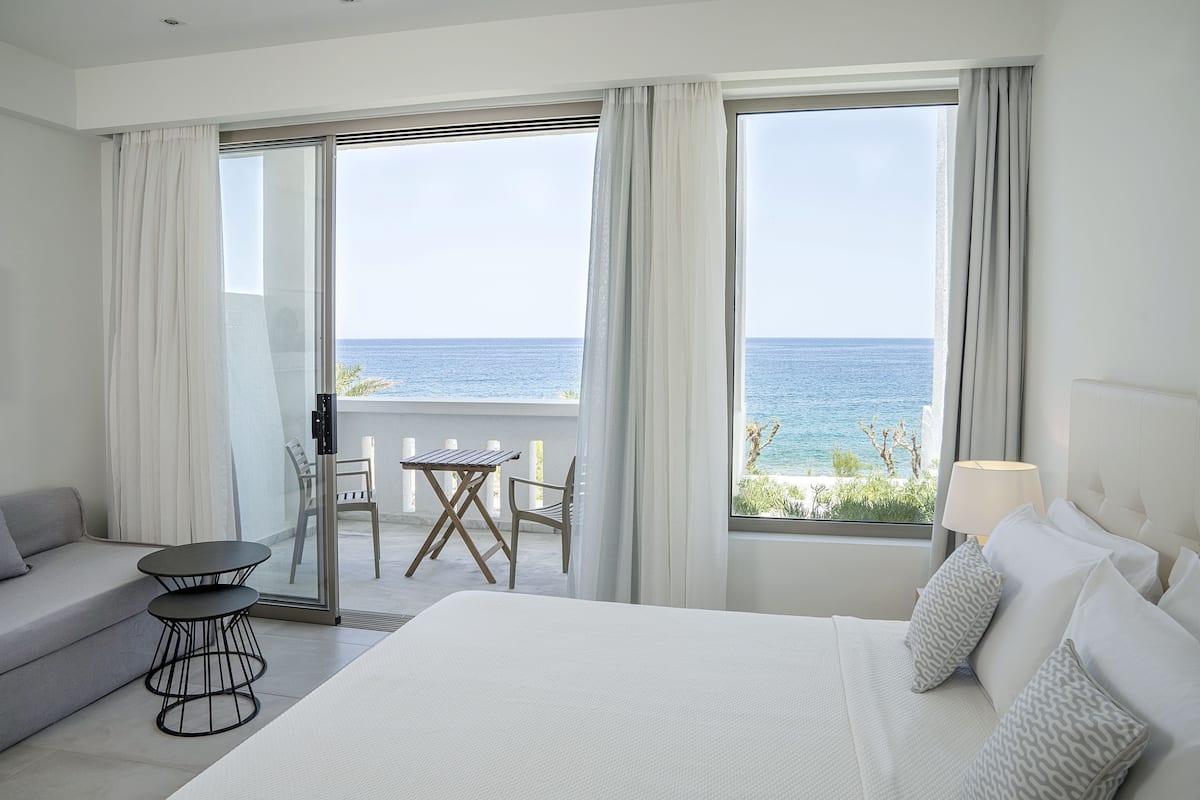 Habitación de hotel con una cama grande, un sofá, una mesita y una silla. Amplias ventanas con cortinas, con vistas al mar.