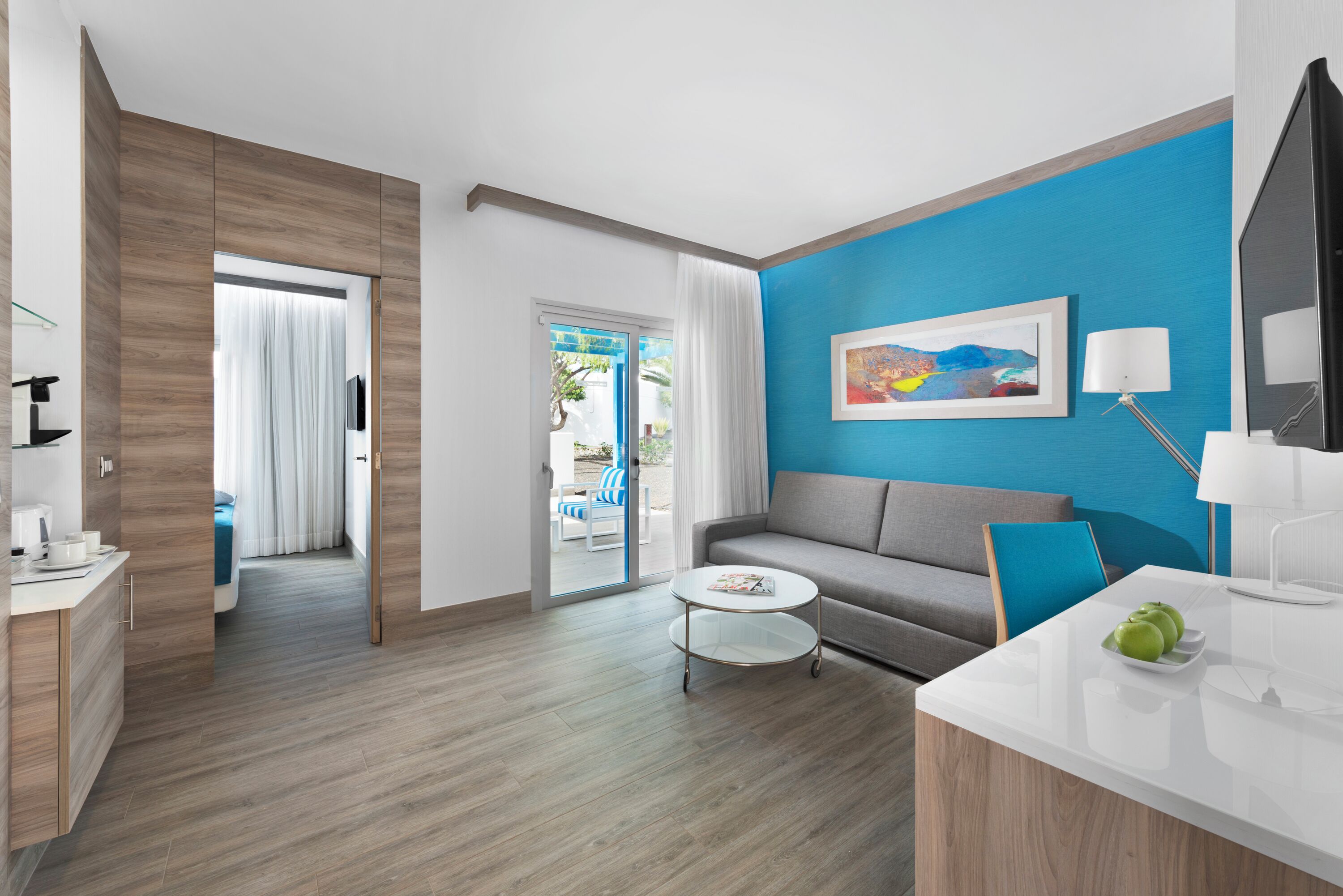 A modern hotel room with a wooden floor, a grey sofa, a white coffee table, and a blue accent wall.