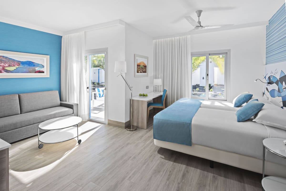 Une chambre d’hôtel moderne avec un canapé gris, un lit avec une literie bleue, un bureau avec une chaise et un ventilateur de plafond.