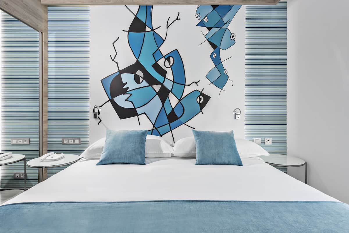 Une chambre d’hôtel moderne avec un grand tableau abstrait au mur, un lit soigneusement fait avec des oreillers bleus et une petite table de chevet avec une lampe.