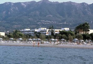 A beach with umbrellas and lounge chairs, a building with multiple floors, and mountains in the background. - Tigaki Beach (Kos)