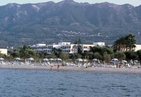 Una playa con sombrillas y sillas de descanso, un edificio de varios pisos y montañas al fondo.