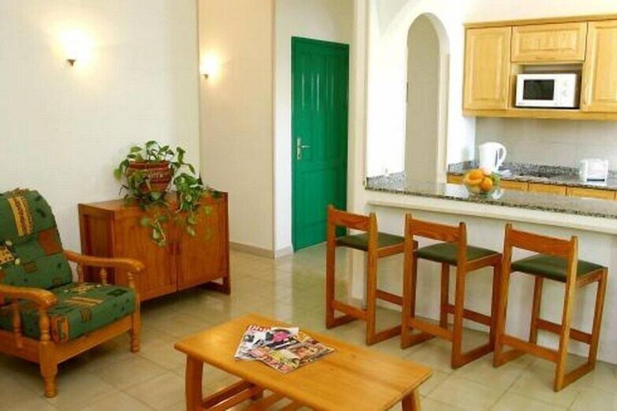 A living room with a green armchair, wooden chairs, a coffee table, and a kitchen area with a microwave and a fruit bowl.