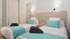 Habitación de hotel con dos camas, cada una con colcha blanca y azul verdoso, y cabeceros de madera. Hay dos lámparas de pared a cada lado de los cabeceros.