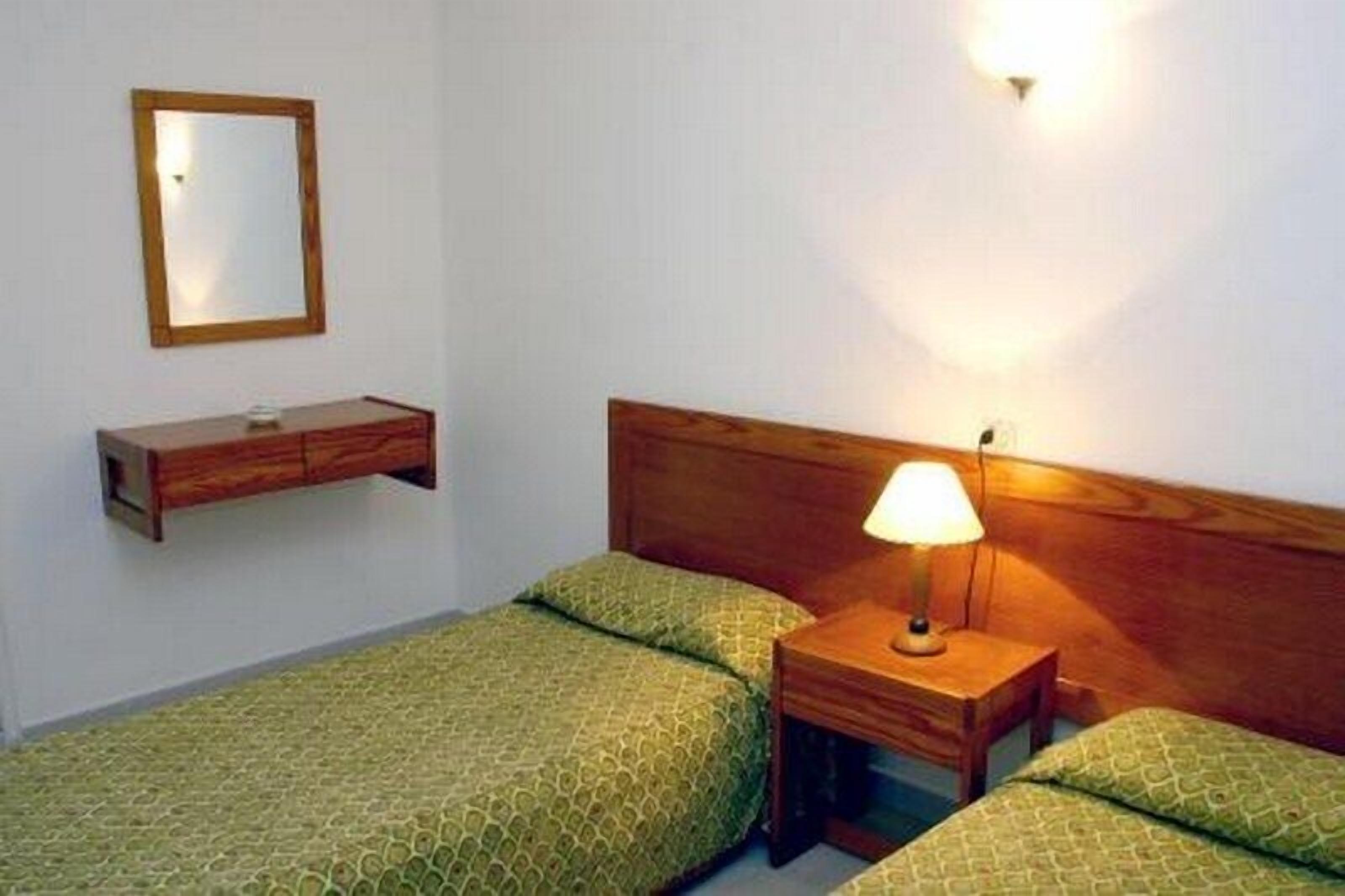 Um quarto pequeno e bem arrumado com duas camas de solteiro, uma mesa de cabeceira com abajur, um criado-mudo de madeira e uma prateleira fixada na parede.