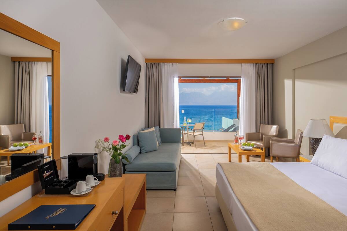 A hotel room with a bed, a sofa, a desk with a coffee maker, a TV, and a balcony with a view of the sea.