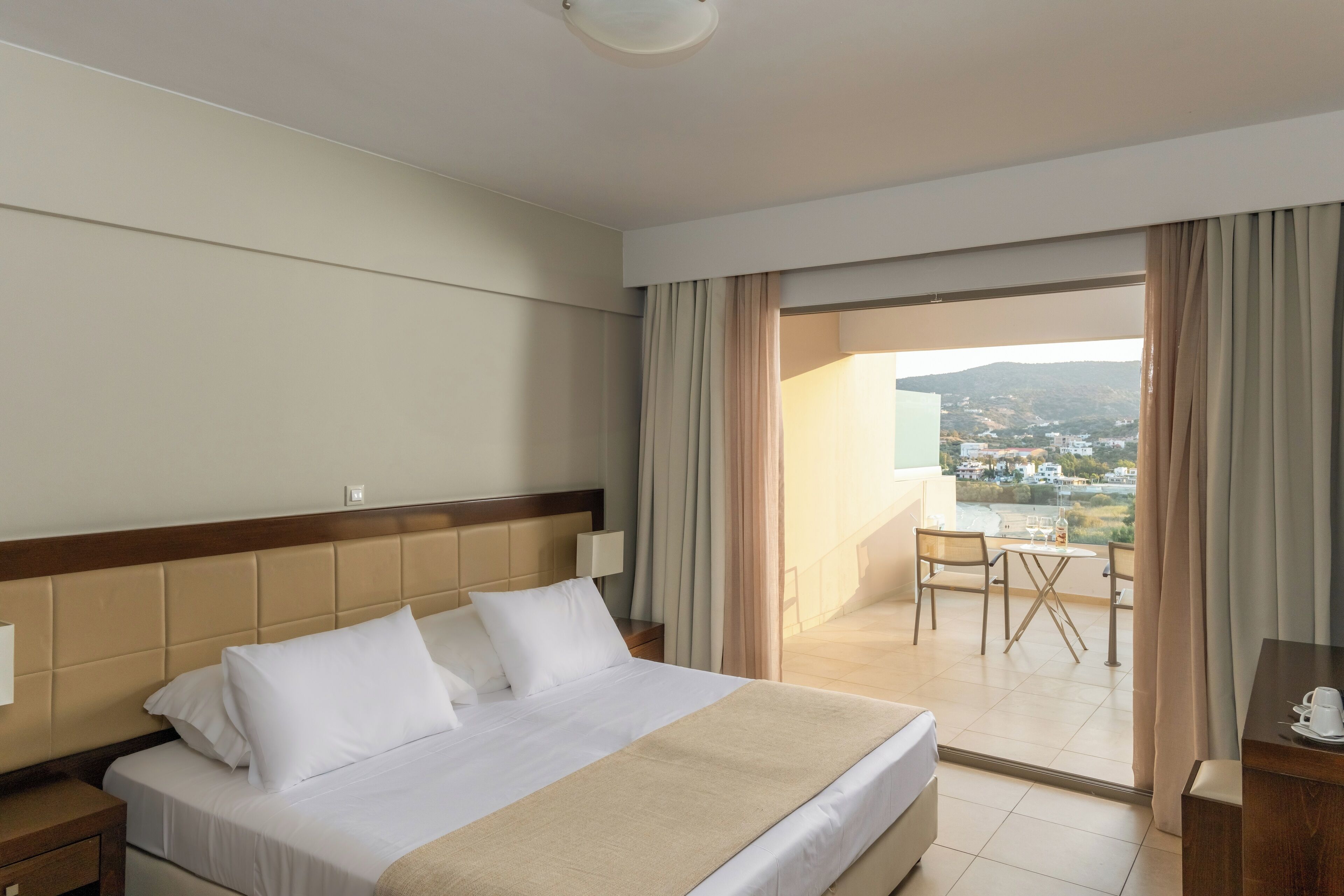 Habitación de hotel con una cama grande, cabecera de madera, mesita de noche, escritorio y balcón con mesa y sillas.