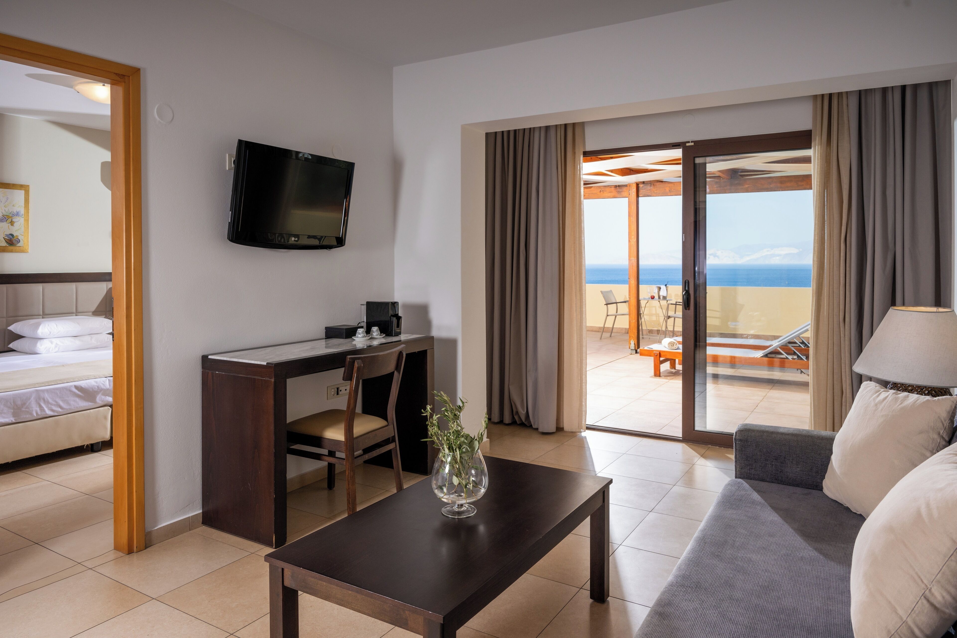 Habitación de hotel con cama, escritorio con silla, televisor, sofá, mesa de centro y balcón con vista al mar.