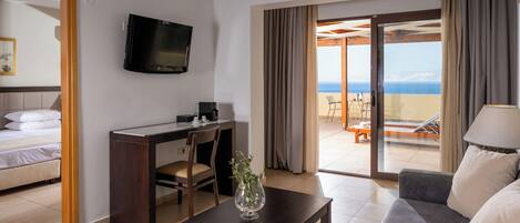 Hotelkamer met een bed, een bureau met stoel, een televisie, een bank, een salontafel en een balkon met uitzicht op zee.