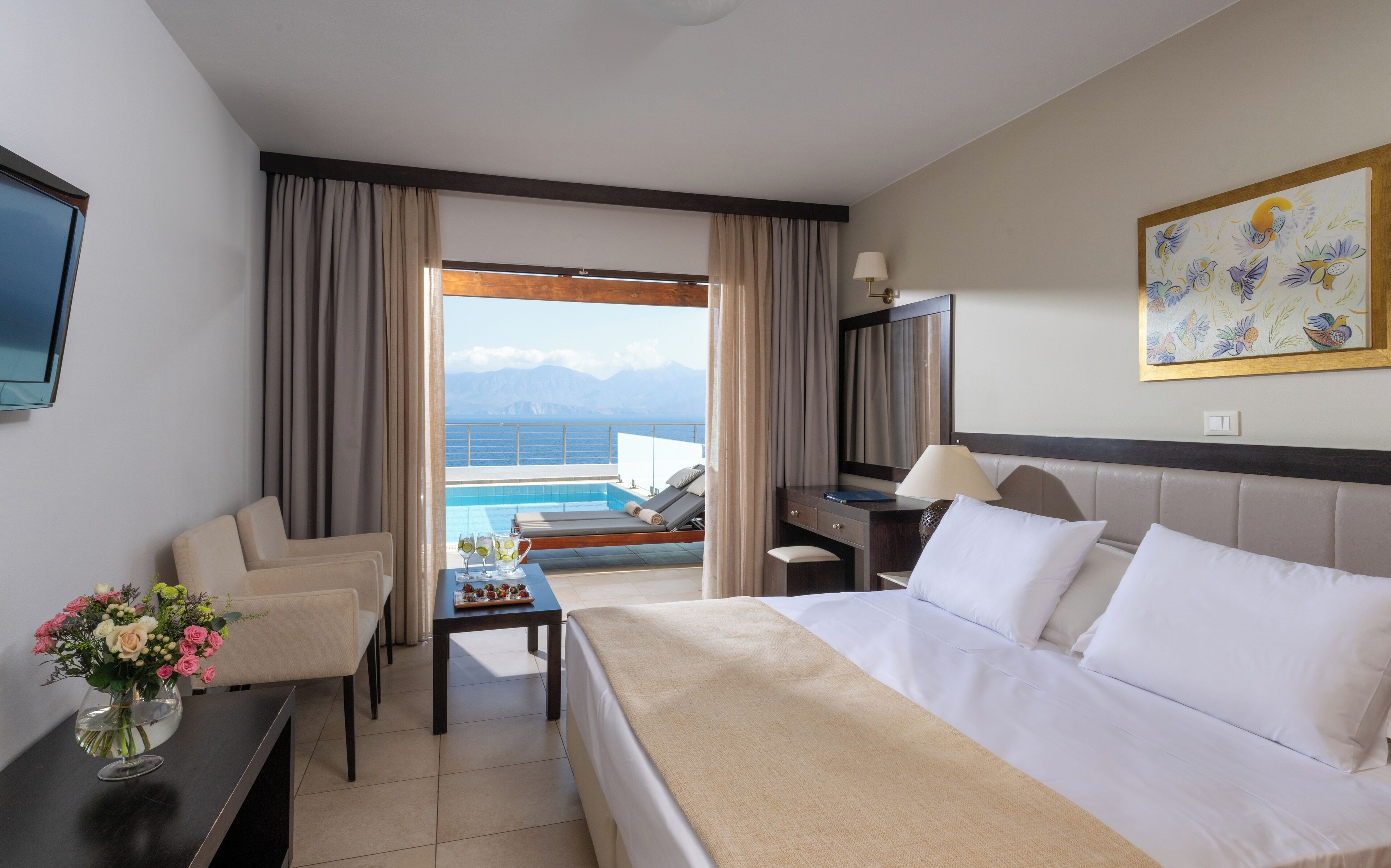 Habitación de hotel con cama, escritorio, televisión, ventana con vista al mar y un cuadro en la pared.