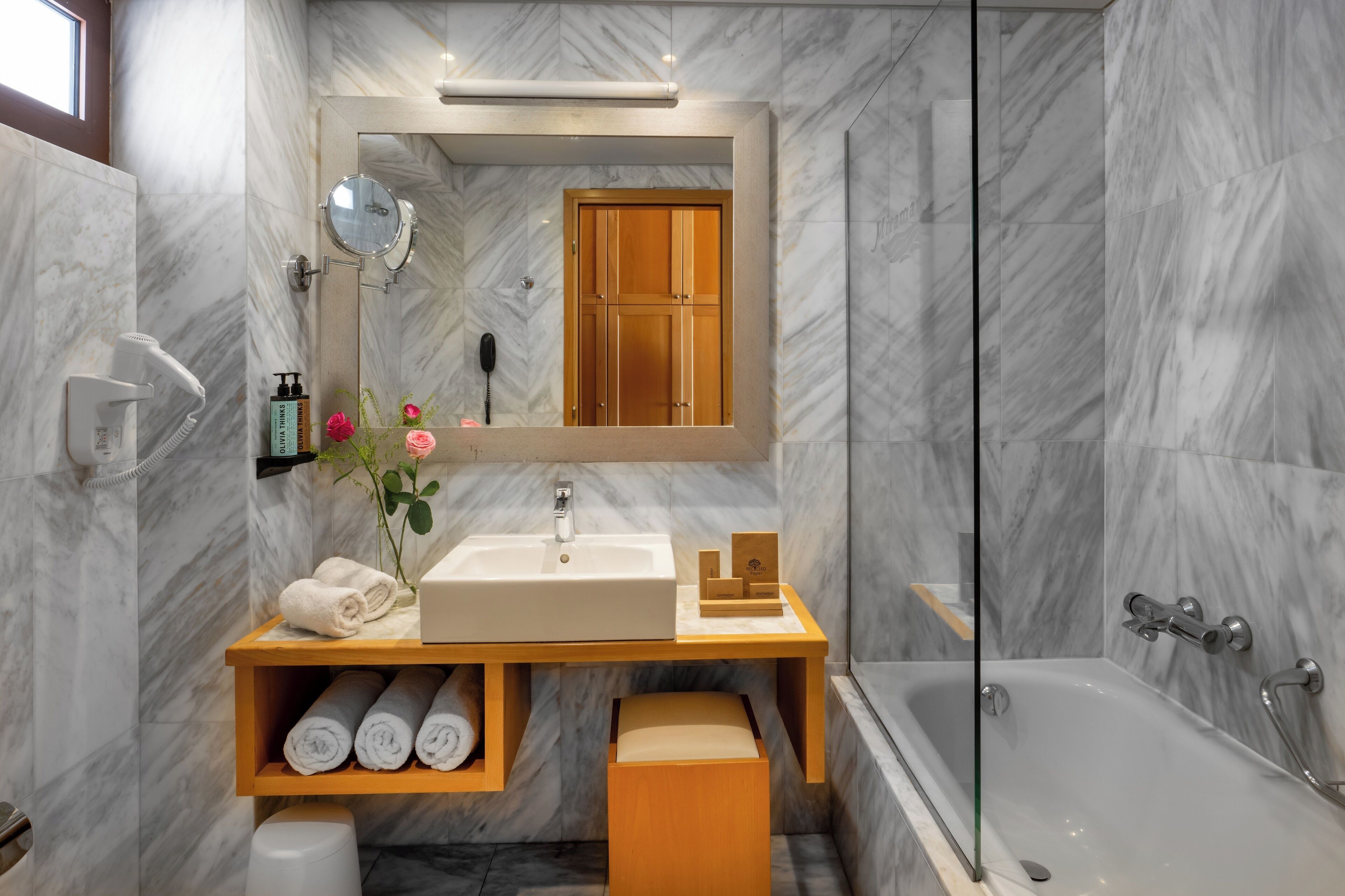 A modern bathroom with a marble wall, a wooden vanity, a sink, a mirror, a towel rack, and a bathtub.