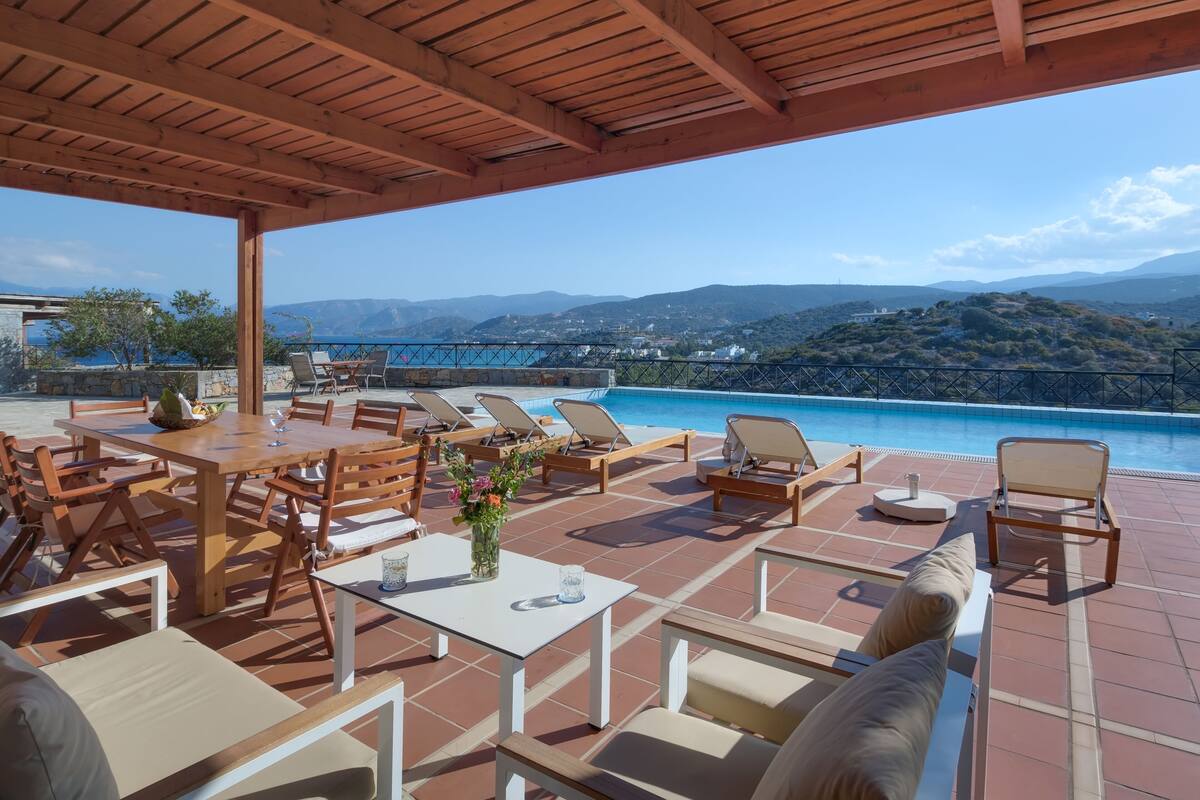 A terrace with wooden furniture, a pool, and a view of mountains and water.