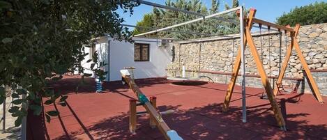 A playground with a seesaw and swings, a stone wall, and a red-tiled floor.