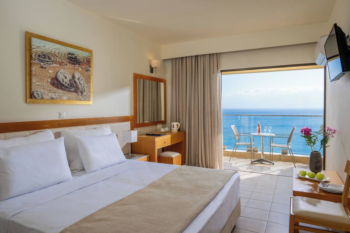 A hotel room with a large bed, a desk, a chair, a small table, and a balcony with a view of the sea.