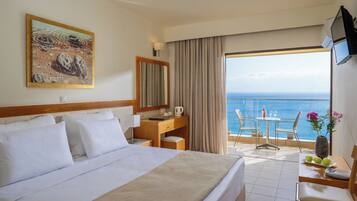 A hotel room with a large bed, a desk, a chair, a small table, and a balcony with a view of the sea.