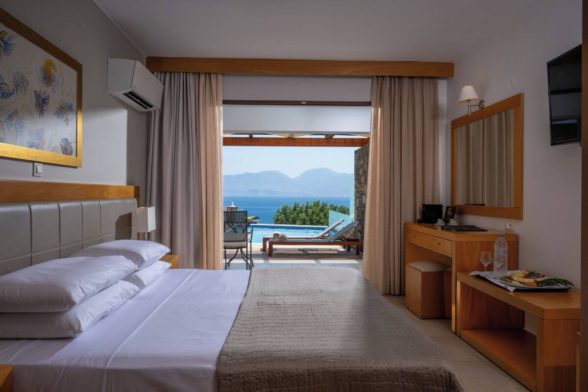 A hotel room with a bed, a desk, a chair, a table, a TV, and a view of the sea and mountains.