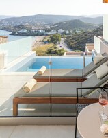 Un área de descanso al aire libre moderna con piscina, barandilla de vidrio y vista a un pueblo costero y colinas.