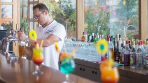 Un barman derrière un comptoir, dans un bar où l’on propose une grande variété de boissons, avec des bouteilles alignées en arrière-plan.