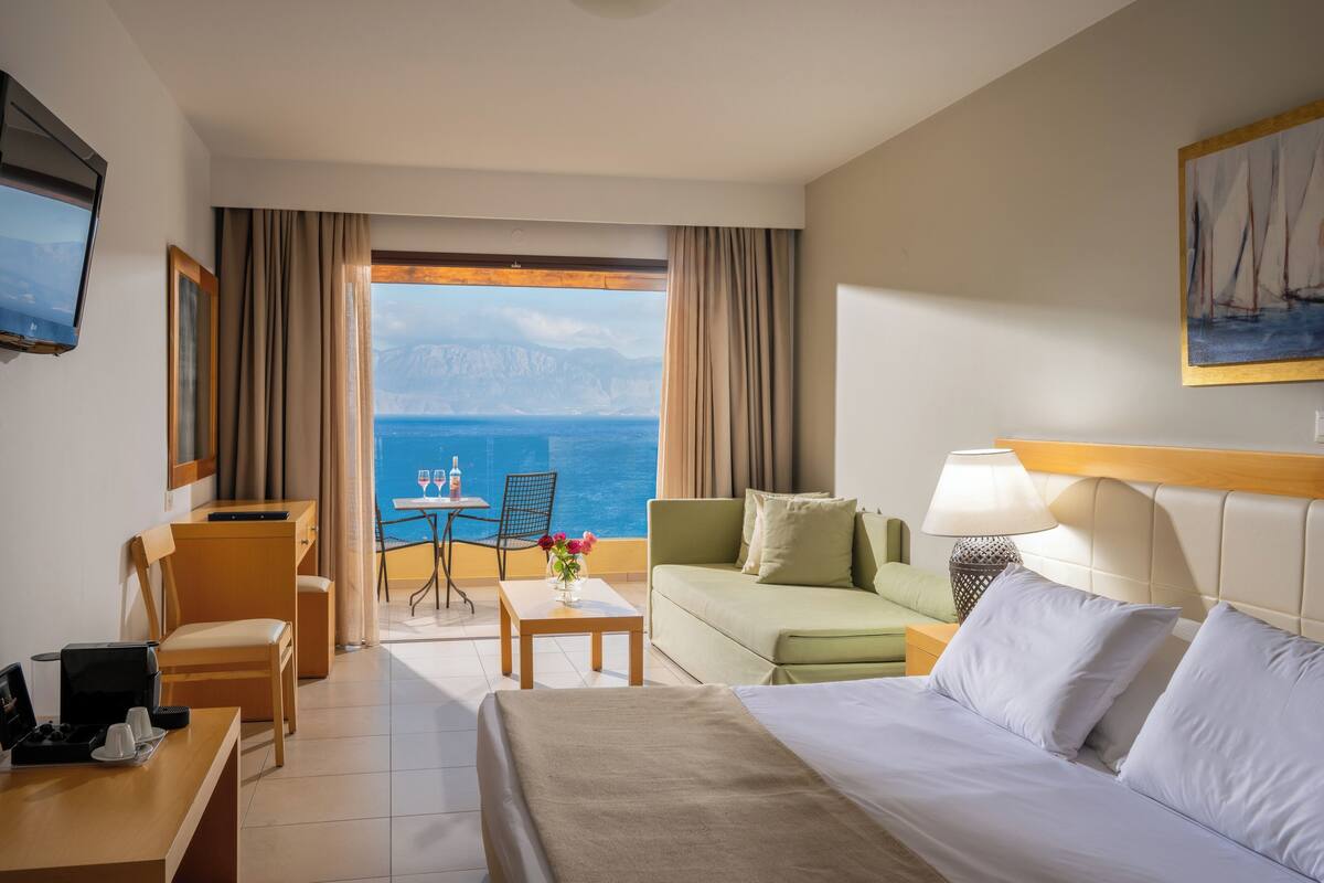 A hotel room with a bed, a sofa, a desk, and a table. There is a view of the sea through the window.