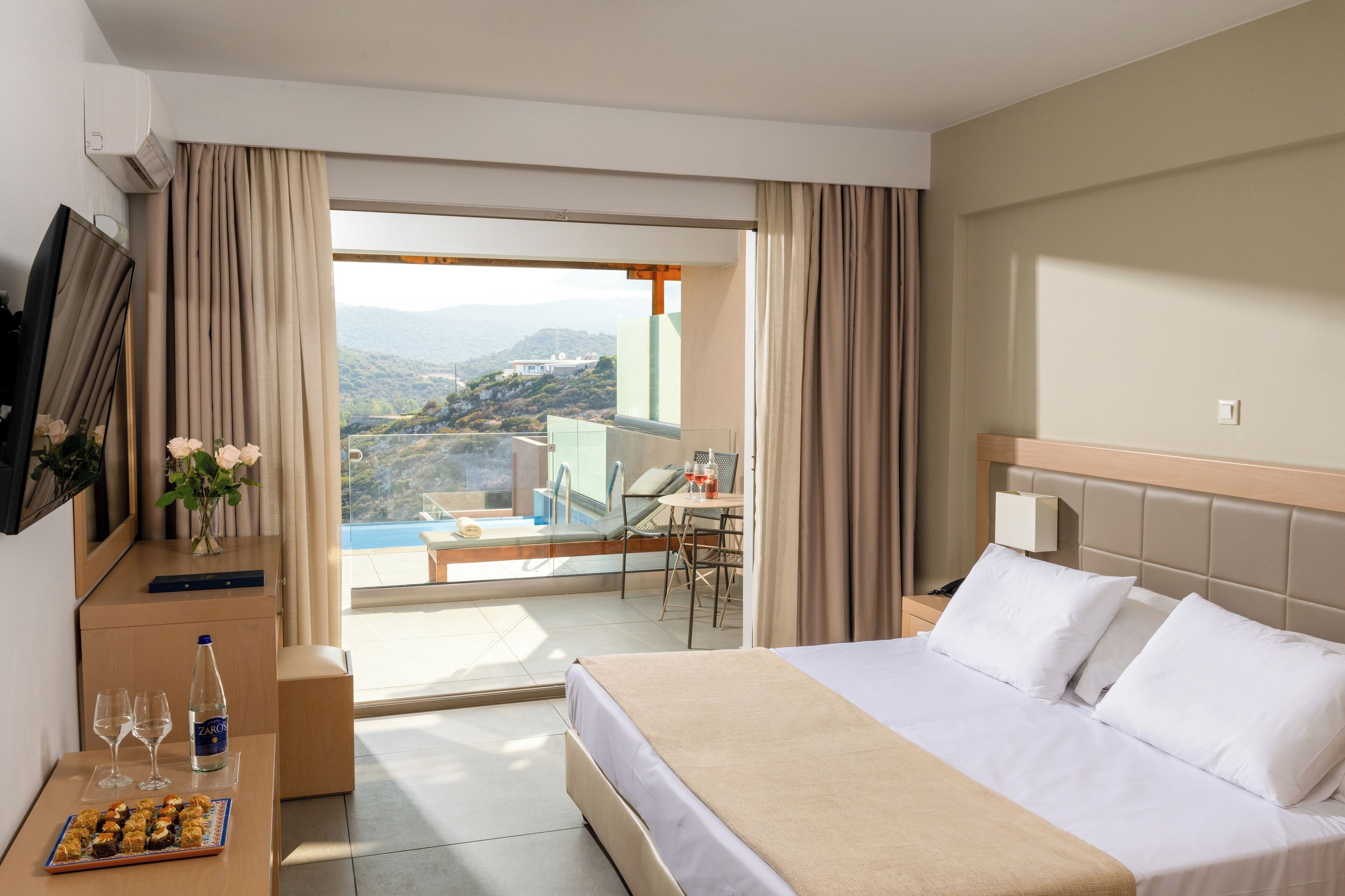 Habitación de hotel con una cama, un escritorio con silla, una mesita con una botella y vasos, y vistas a una piscina y montañas.