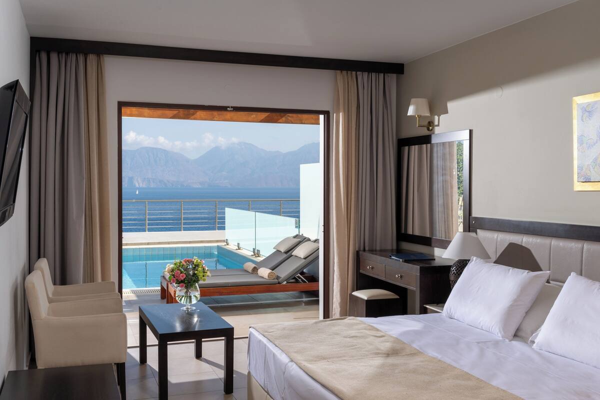 A hotel room with a bed, a desk, a chair, a small table with a vase of flowers, and a view of a pool and mountains.