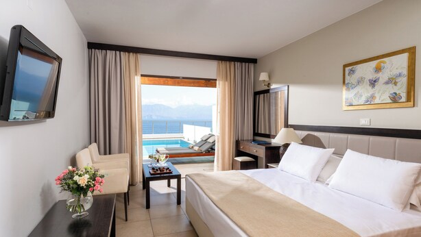 A hotel room with a bed, a desk, a TV, a view of the pool and mountains. - Miramare Resort & Spa (Agios Nikolaos)