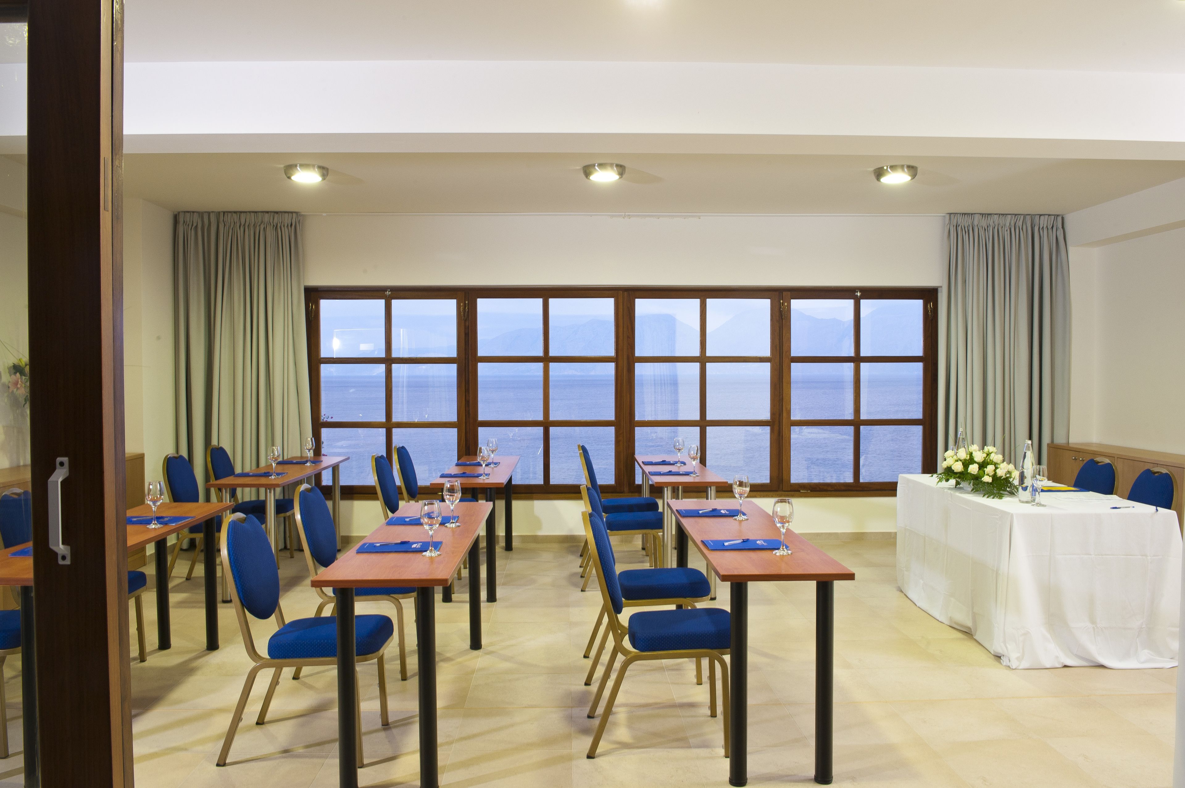 Une salle de conférence équipée de tables et de chaises, de grandes fenêtres offrant une vue sur la mer, et une table blanche décorée de fleurs.