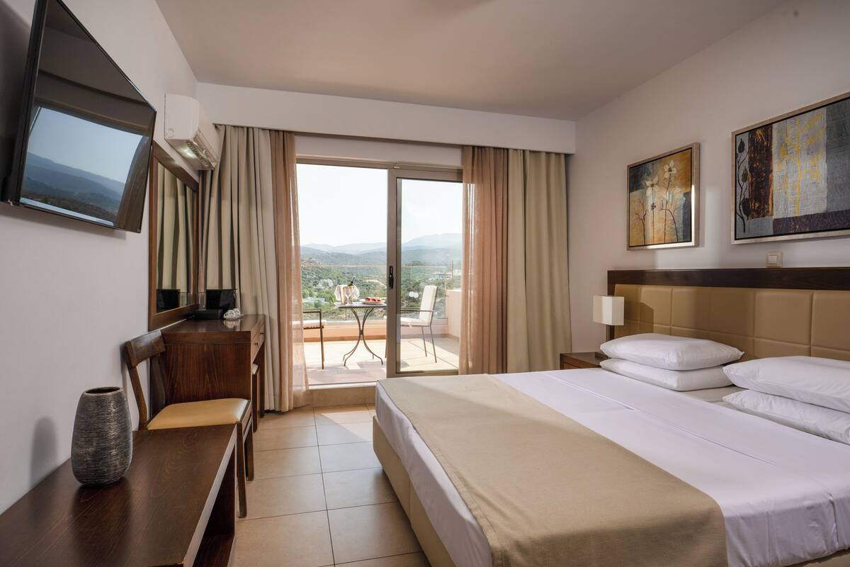 A hotel room with a bed, a desk, a chair, a TV, a painting, and a balcony with a view.