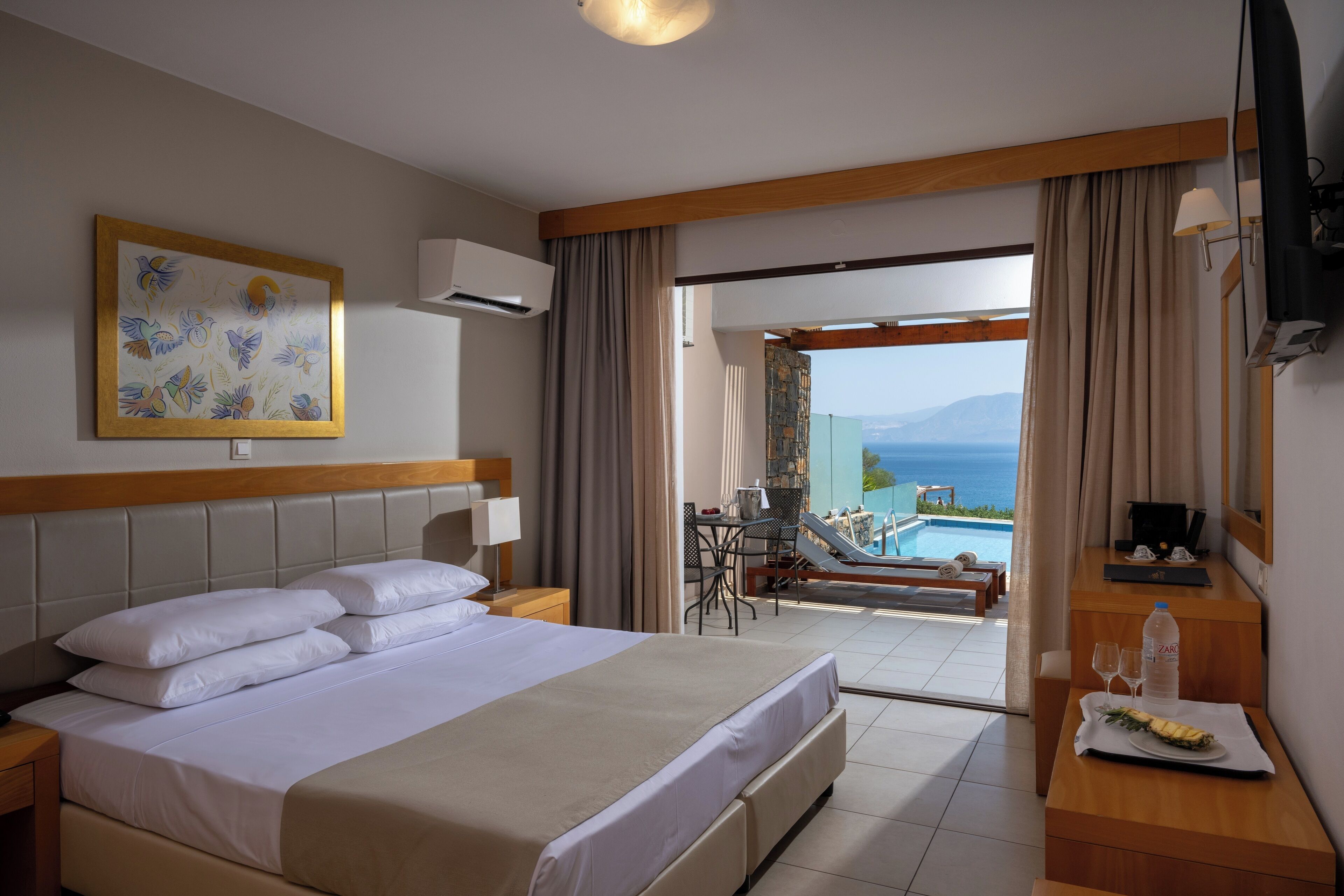 A hotel room with a large bed, a desk with a chair, a dining area with a table and chairs, and a view of the ocean.