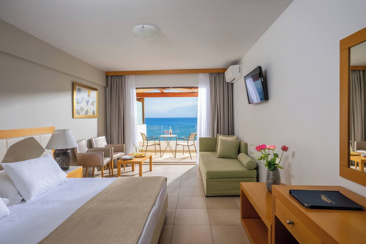 A hotel room with a bed, a sofa, a desk, a TV, a window with a view of the sea, and a vase with flowers.