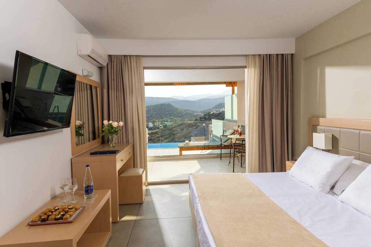 A hotel room with a bed, a TV, a desk with a laptop, a chair, and a view of a mountainous landscape.