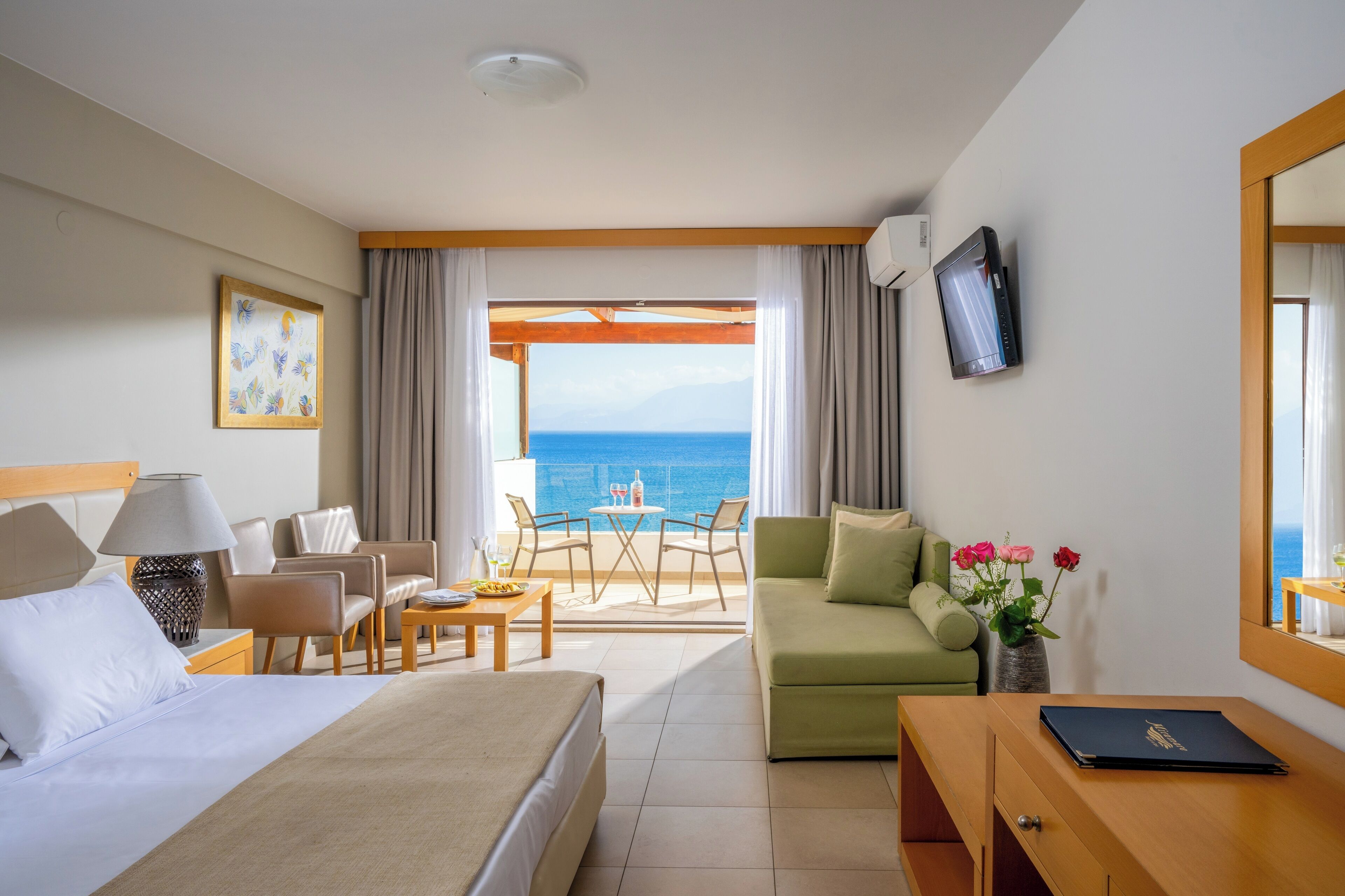 Habitación de hotel con cama, sofá verde, escritorio, televisor y balcón con vista al mar.
