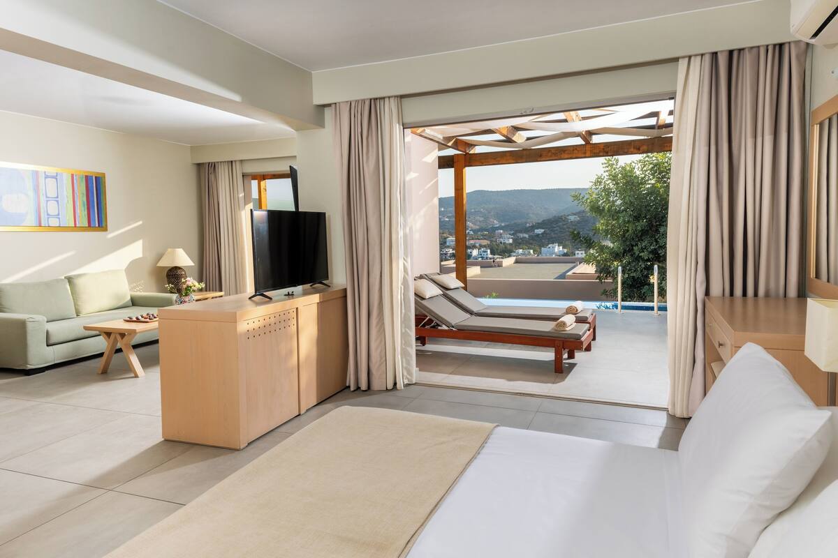 A modern hotel room with a large bed, a desk with a flat-screen TV, a sofa, and a view of a pool and mountains.