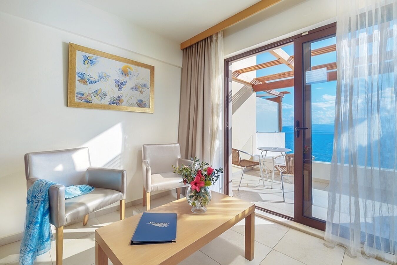 A hotel room with two chairs, a table with a book, a painting, and a view of the sea through a glass door.
