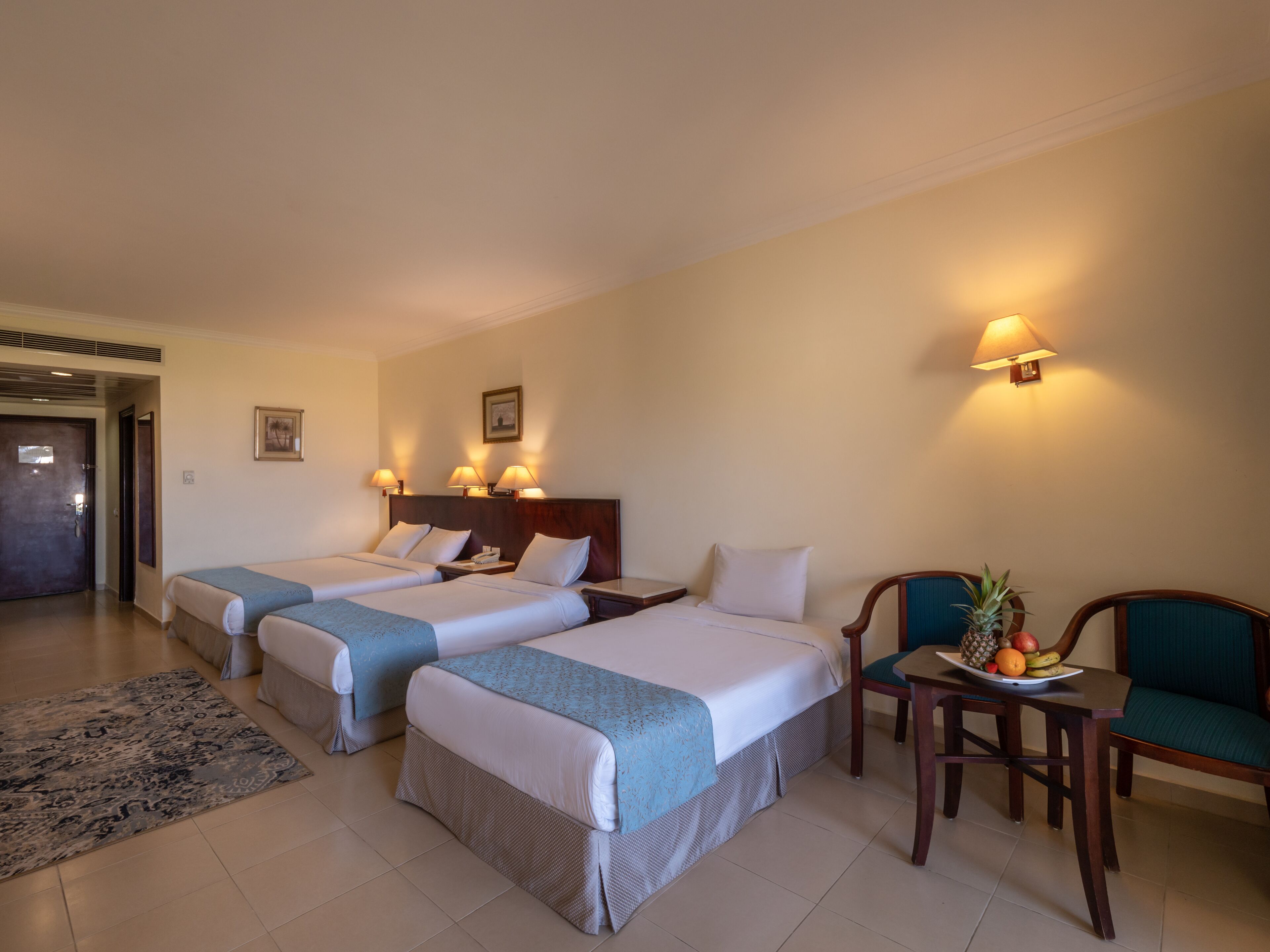 A hotel room with two beds, a small dining table with fruit, a chair, and a door leading to a hallway.