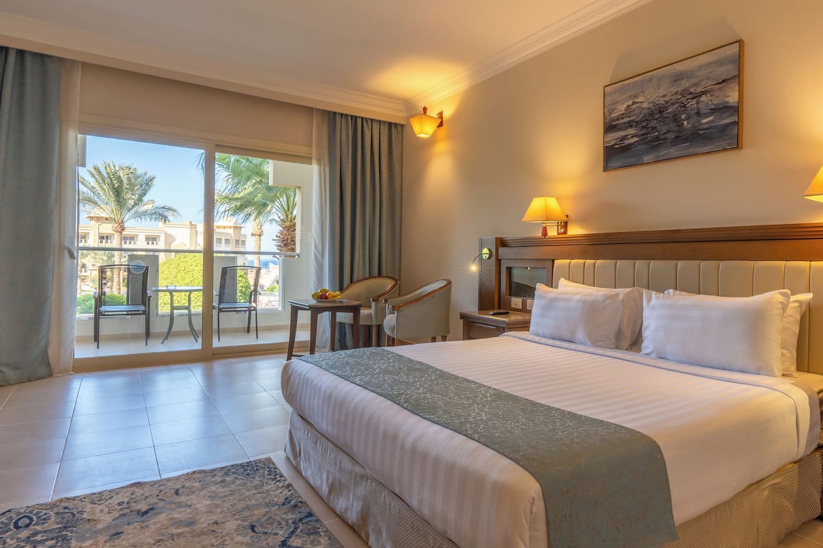 A hotel room with a bed, two bedside lamps, a painting, a chair, a table, and a view of palm trees through the balcony doors.