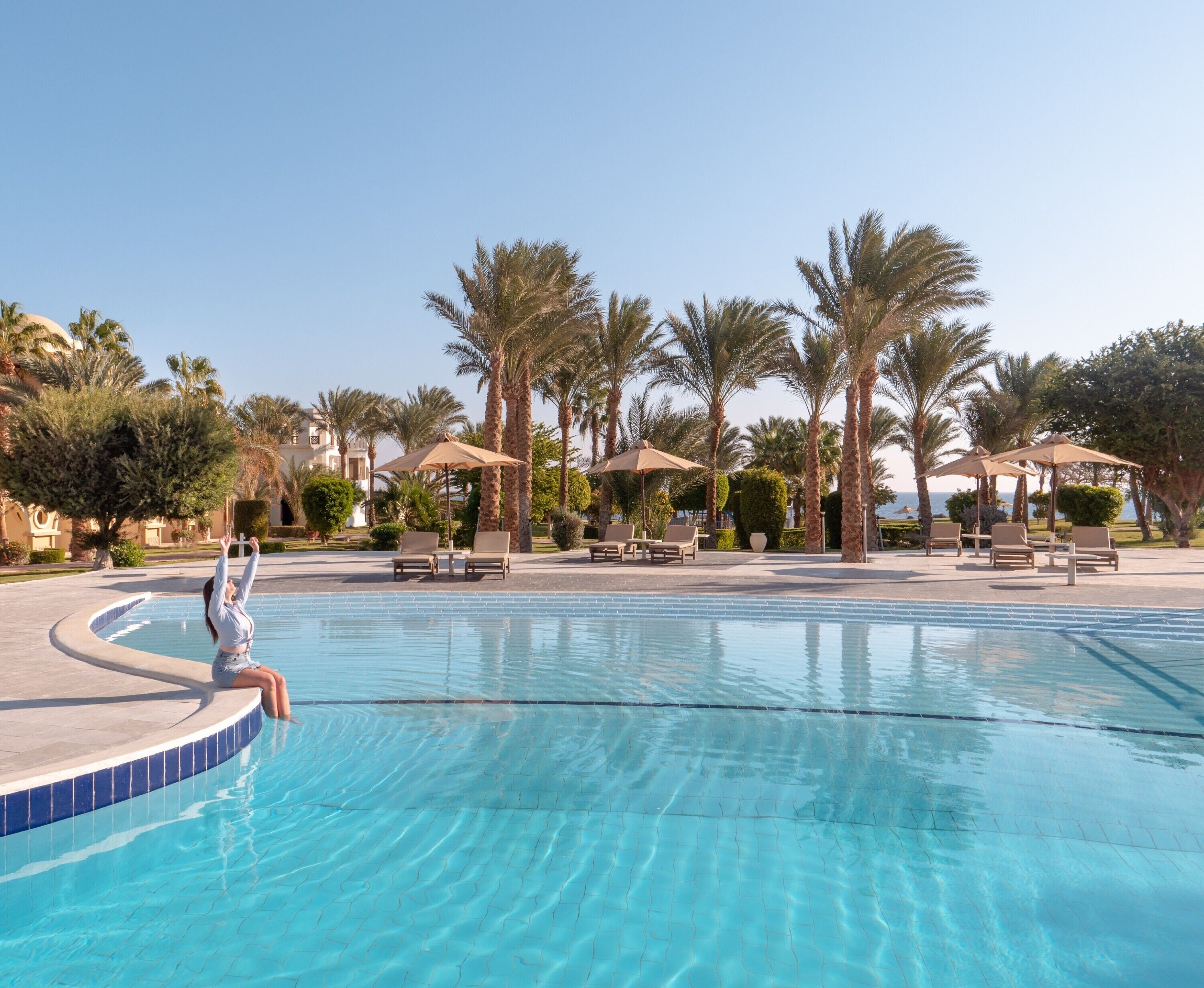 A swimming pool with lounge chairs and umbrellas, surrounded by palm trees and a clear sky.