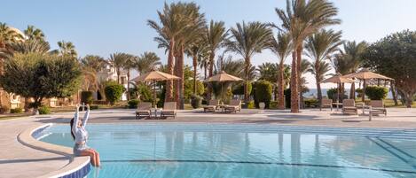 A swimming pool with lounge chairs and umbrellas, surrounded by palm trees and a clear sky.