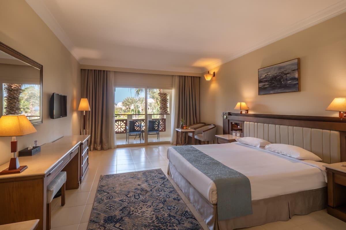 A hotel room with a large bed, a desk, a chair, a TV, and a balcony with a view of palm trees.