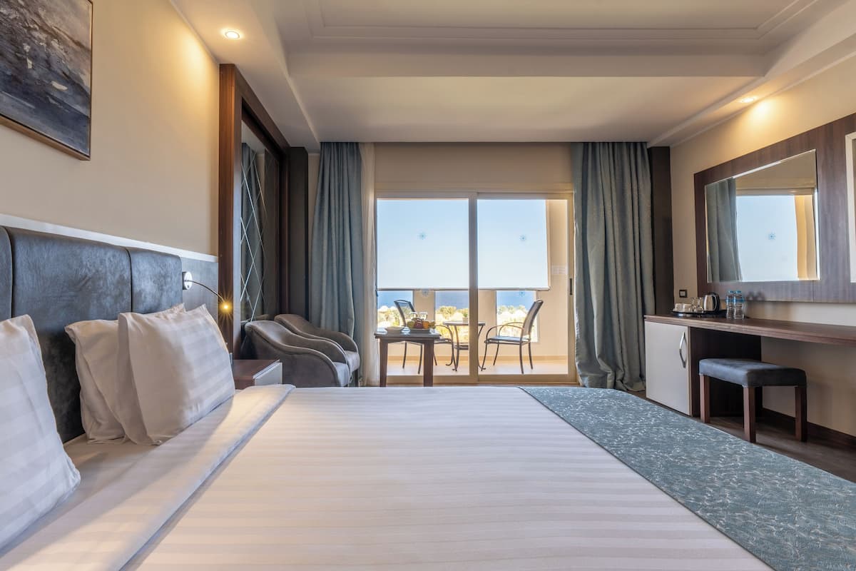 A hotel room with a large bed, a desk with a chair, a small table with a chair, and a balcony with a view of the sea.