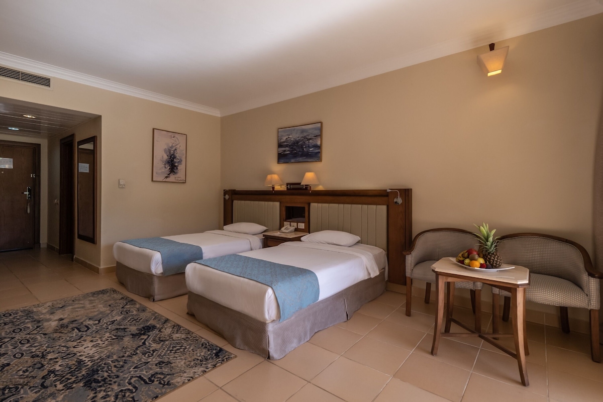 Habitación de hotel con dos camas, una mesita con fruta, una silla, una alfombra y obras de arte en las paredes.