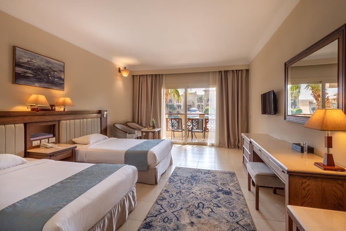 A hotel room with two beds, a desk, a chair, a TV, and a balcony with outdoor furniture.
